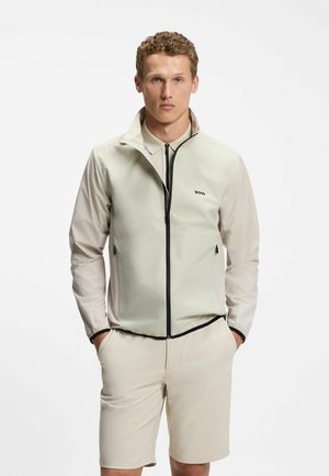 Mand iført beige lynlåsvest over matchende langærmet skjorte og shorts, stående med hænderne i lommerne mod hvid baggrund.
