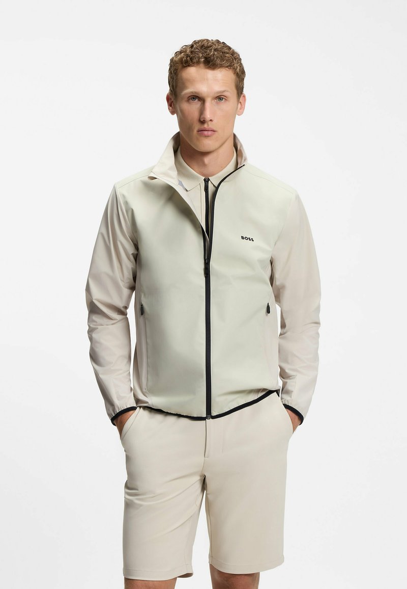 Homme portant un gilet beige zippé par-dessus une chemise à manches longues assortie et un short, debout les mains dans les poches devant un fond blanc.