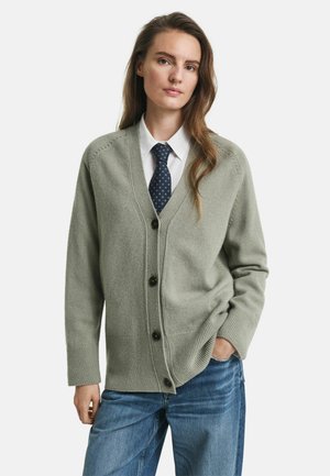 Cardigan vert clair en tissu doux, avec un col en V, une fermeture à boutons et des poignets côtelés, associé à un jean en denim et une chemise blanche.