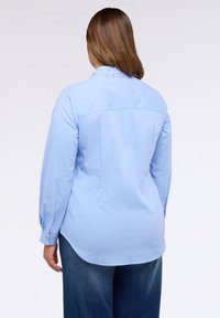 Donna con capelli castani che indossa una camicia a maniche lunghe a righe verticali blu chiaro con decorazioni di perle sul colletto, abbinata a jeans blu scuro.