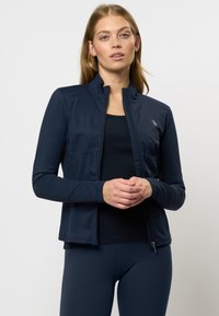 Vrouw in een marineblauwe ritsjack en bijpassende legging, met handen gevouwen en een neutrale uitdrukking tegen een effen achtergrond.