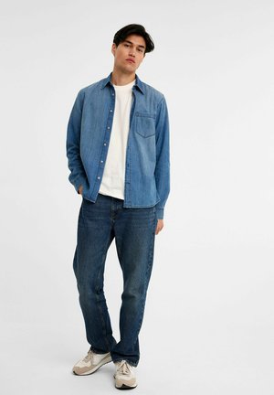 Ljusblå denimskjorta med bröstficka, bärs över en vit t-shirt och matchas med mörkblå jeans. Inkluderar vardagliga sportskor.