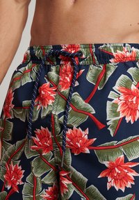 Shorts da nuoto blu navy con un vivace motivo floreale rosso e verde, con una vita elasticizzata e un cordoncino blu scuro con accenti bianchi.