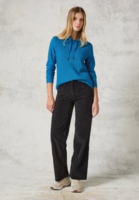 Pull en maille bleue avec cordons, associé à un pantalon en velours côtelé noir et des baskets beiges, sur un fond clair texturé.