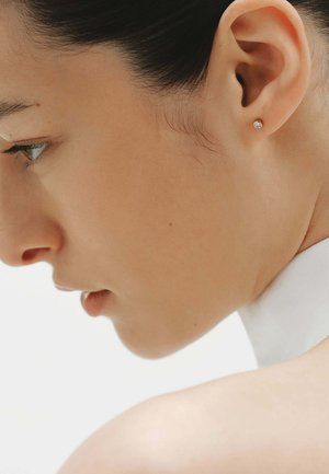 Joven en perfil con piel suave, cabello oscuro recogido hacia atrás, lleva un pequeño pendiente de botón y una prenda blanca de cuello alto.