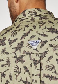 Groene vissersshirt met een camouflagepatroon van vissen, haken en aquatische vormen; bevat een driehoekig PFG-label op de achterkant.