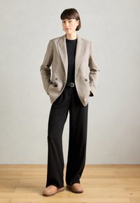 Blazer s houndstooth vzorom s dvoma prednými gombíkmi, čierny top, čierne široké nohavice a béžové slip-on topánky na drevenej podlahe.