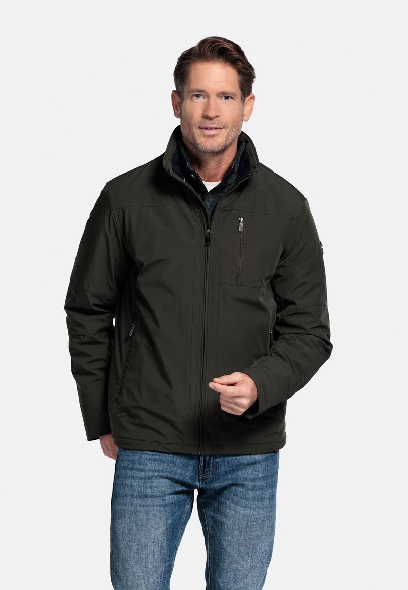 Donkergroene geïsoleerde jas met een hoge kraag, een ritszak aan de voorkant en zijzakken. Draagt over een marineblauw shirt, weergegeven tegen een effen achtergrond.