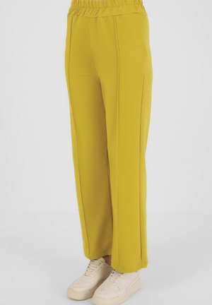 Pantalon ample jaune moutarde avec une ceinture élastique porté avec des baskets blanches à plateforme sur un fond uni.