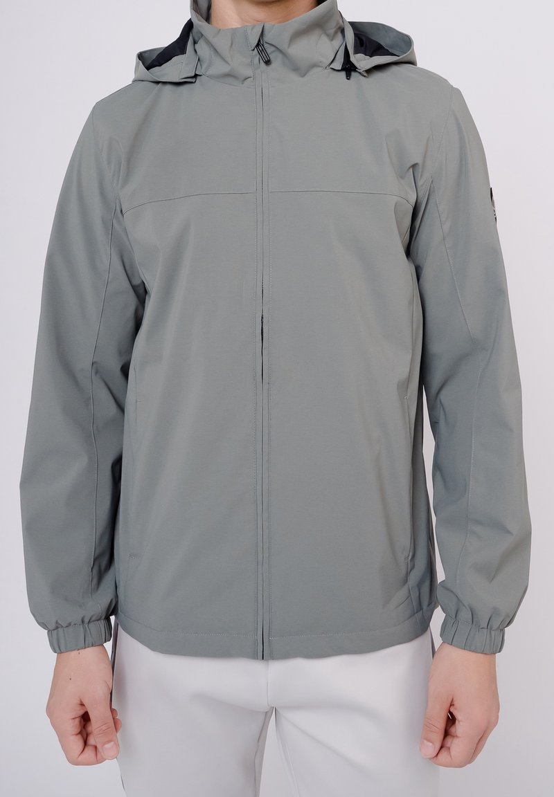 Homme portant une veste à capuche gris clair avec fermeture éclair sur le devant et des poignets élastiques, associée à un pantalon blanc, debout devant un fond uni.