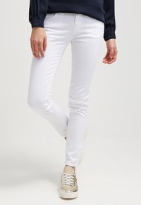 Vita smala jeans med en slät textur, framfickor och en midjehöjd som ligger mitt emellan, i kombination med ljusa sneakers med metalliska detaljer.