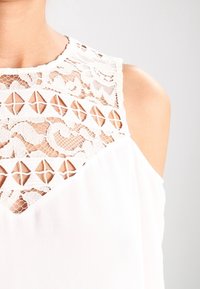 Blouse blanche avec détails en dentelle comprenant des découpes géométriques sur les épaules et le décolleté, texture de tissu lisse, et coupe décontractée.