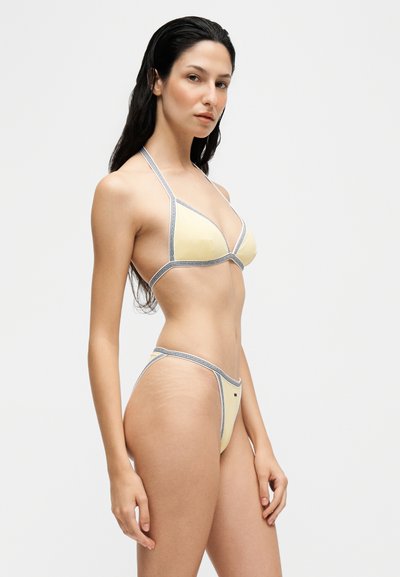 Jeune femme aux longs cheveux foncés portant un bikini jaune pâle avec un liseré contrastant, debout devant un fond clair uni.