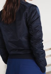 Giacca bomber marina con una finitura lucida e liscia. Presenta polsini e orlo a coste, insieme a dettagli arricciati sulle spalle.