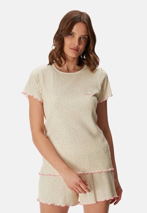 Femme aux cheveux châtains mi-longs portant un top crème à manches courtes et un short avec de petits pois colorés et des bordures froufroutantes roses.