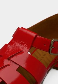 Sandale en cuir verni rouge avec des larges brides, une fermeture à boucle et une semelle texturée, avec un design à bout ouvert.