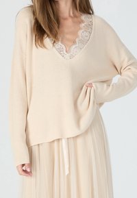 Pull beige en tricot avec un décolleté en V profond, orné d'une bordure en dentelle. Les manches sont longues et larges, associées à une jupe superposée et fluide.