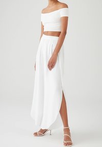 Witte off-shoulder crop top gecombineerd met een lange witte geplooide rok met een zijsplit, een gladde textuur en strappy hoge hakken.