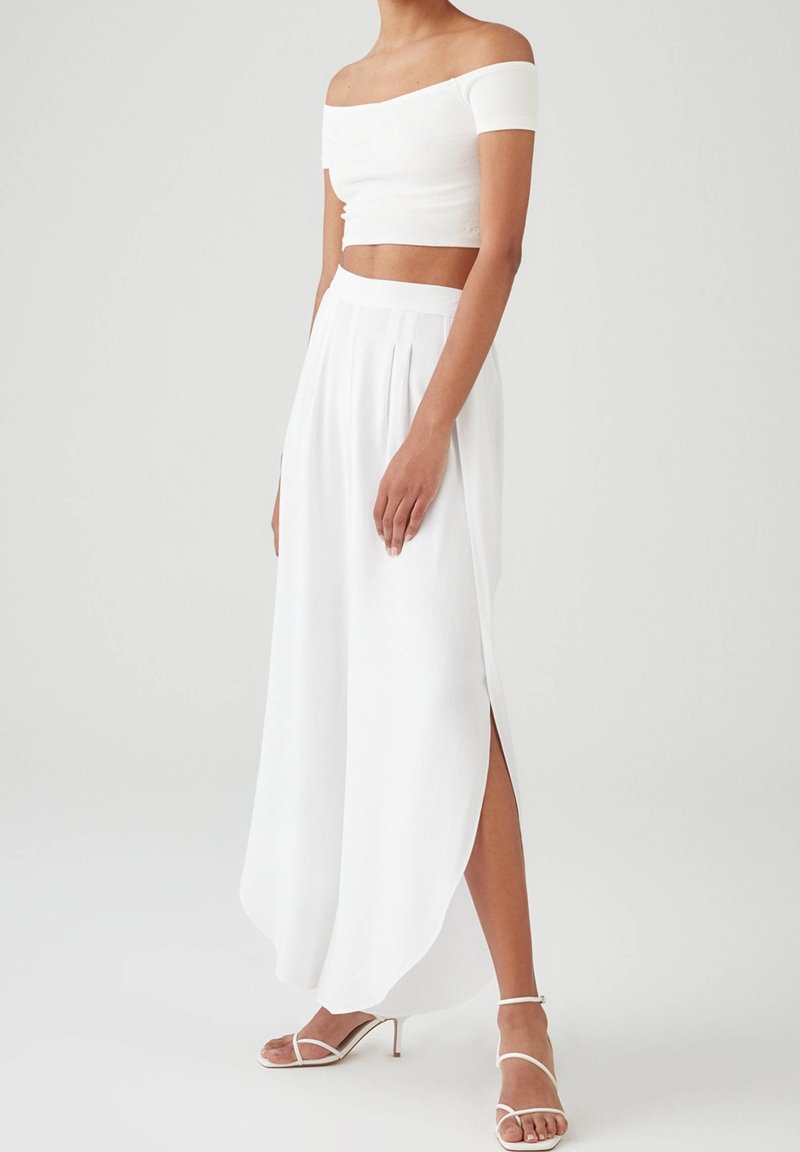 Witte off-shoulder crop top gecombineerd met een lange witte geplooide rok met een zijsplit, een gladde textuur en strappy hoge hakken.