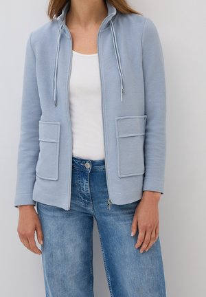 Frau trägt eine hellblaue Jacke mit Reißverschluss und großen Fronttaschen, ein weißes Oberteil und blaue Jeans, steht vor einem schlichten Hintergrund.