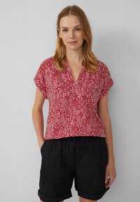 Blouse à manches courtes rouge à motif, avec un col en V, présentant un design géométrique en blanc. Associée à un short noir à revers.