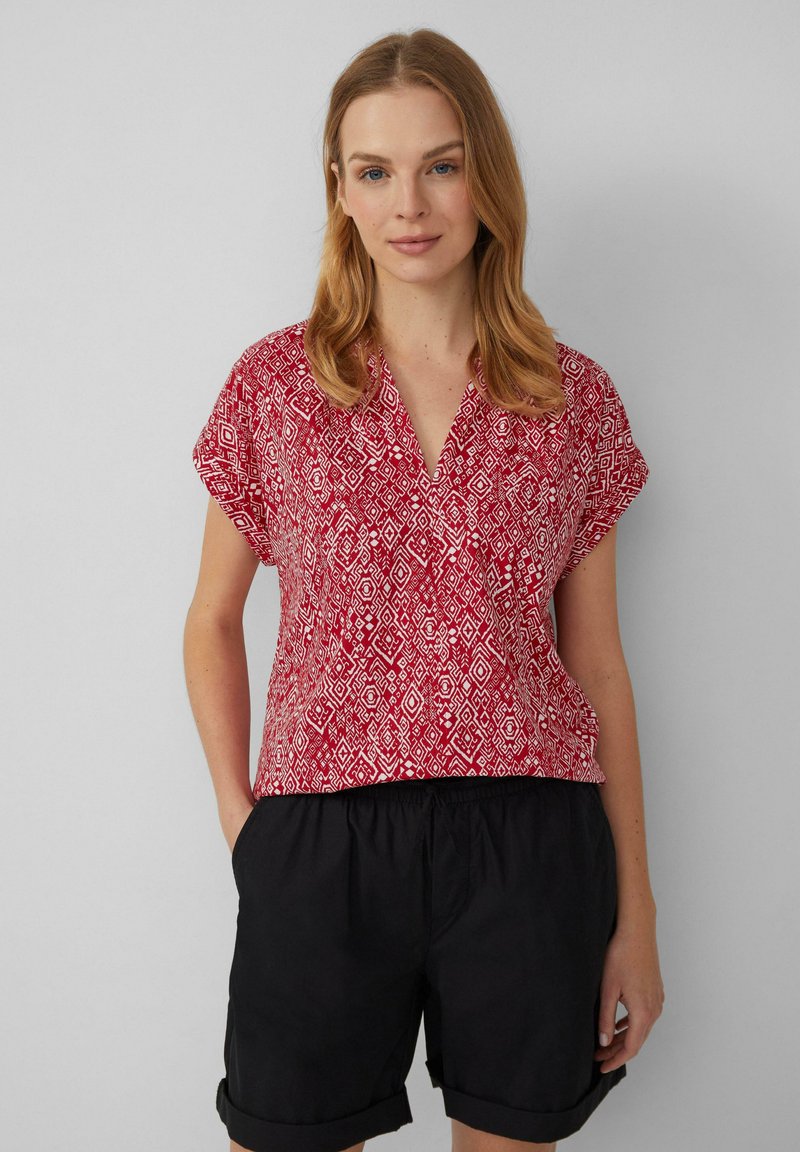 Blouse à manches courtes rouge à motif, avec un col en V, présentant un design géométrique en blanc. Associée à un short noir à revers.