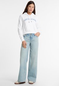 Hoodie bianco a collo corto con la scritta "Guess Jeans" stampata in blu, abbinato a jeans a gamba larga blu chiaro. La calzatura presenta accenti beige e marroni.