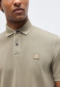 Άνδρας μοντέλο που φοράει ελαφρώς ελιά πράσινο πόλο με δύο σκούρα κουμπιά και ένα μικρό τετράγωνο λογότυπο στο αριστερό στήθος.