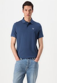 Tumedast sinine polo särk, mis on valmistatud pehmest puuvillast, sisaldab kraed ja lühikesi varukaid. Rinna peal on diskreetne brodööritud logo.