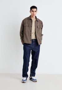 Brown fleece shirt jacket with two chest pockets, beige t-shirt underneath, dark blue denim pants, and blue/white sneakers with gray accents.
