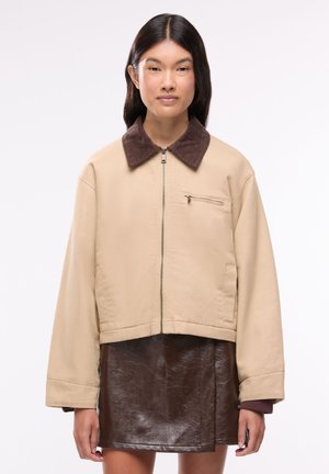 Giovane donna che indossa una giacca beige con colletto marrone e una gonna in pelle marrone scuro, in piedi davanti a uno sfondo bianco uniforme.