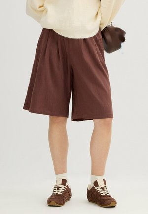 Persona con pantalones cortos marrones de pierna ancha, calcetas blancas, zapatillas marrones, suéter crema y sosteniendo un bolso marrón sobre un fondo liso.