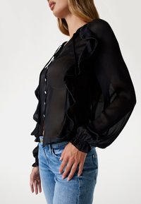 Svart genomskinlig blus med volangdetaljer framtill, knappstängning och elastiska manschetter. Kombineras med blå jeans.