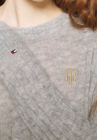 Pull gris tricoté présentant un motif texturé, un design en câble et un logo brodé en or. Petite pièce tricolore sur la manche.