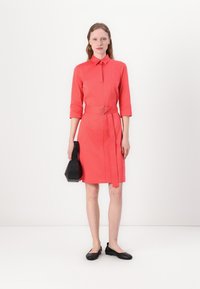 Coral shirt dress with a collared neckline, three-quarter sleeves, belt detail, and a side pleat. Worn with black flats and a small black handbag.