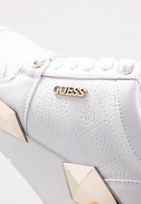 Weiße Sneakers mit eingeprägtem Logo-Muster, goldenen Hardware-Akzenten, strukturiertem Obermaterial und dreieckigen goldenen Nieten an der Seite.