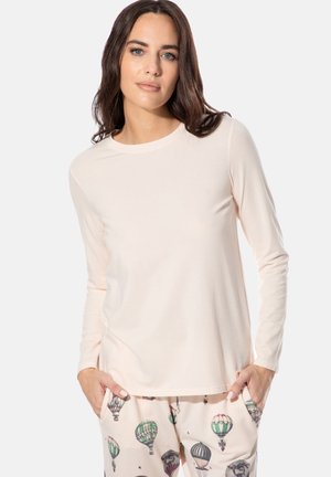 Frau trägt ein hellbeiges langärmeliges Hemd und cremefarbene Hosen mit bunten Heißluftballon-Motiven, Hände in den Taschen, neutraler Hintergrund.