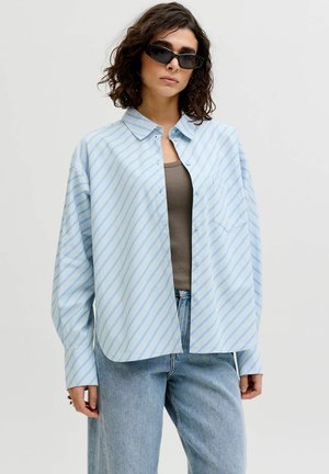 Frau mit lockigem Haar, die schwarze Sonnenbrillen trägt, ein übergroßes hellblau gestreiftes Hemd über einem braunen Top und hellblaue Jeans vor einem einfarbigen Hintergrund.