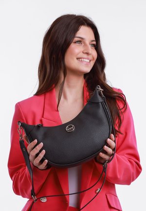 Lächelnde Frau mit langen braunen Haaren, die ein rotes Blazer trägt, hält eine strukturierte schwarze, halbmondförmige Handtasche mit silbernem Logo und Trageriemen.