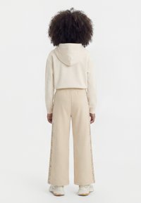 Παιδί με σγουρά μαλλιά στέκεται με την πλάτη, φοράει ένα μπεζ φούτερ με κουκούλα και αντίστοιχο παντελόνι με πλατύ πόδι και ρίγες με σχέδια στα πλαϊνά.