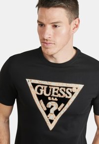 Camiseta negra de manga corta con un logotipo triangular bordado en oro y azul, con patrones detallados y texto dentro del triángulo.