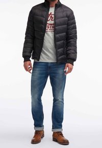 Giacca a vento nera con design trapuntato, t-shirt grigia a girocollo con testo, jeans di denim blu e scarponi in pelle marrone con risvolti.