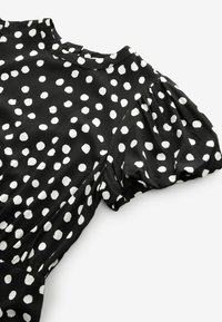 Svart blus med vita prickar, korta puffärmar, rundad halsringning och texturerad tyg. Visuellt distinkt lekfullt mönster.