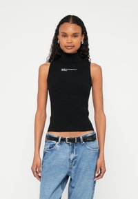 Γυναίκα με σγουρά μαύρα μαλλιά που φοράει μαύρο αμάνικο τοπ με ψηλό λαιμό και φαρδιά μπλε τζιν με μαύρη ζώνη, στέκεται μπροστά σε λευκό φόντο.