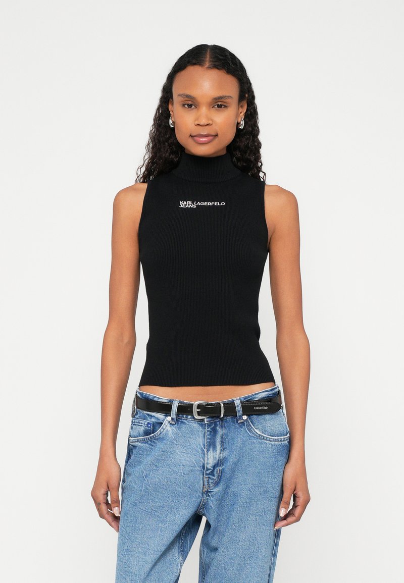 Γυναίκα με σγουρά μαύρα μαλλιά που φοράει μαύρο αμάνικο τοπ με ψηλό λαιμό και φαρδιά μπλε τζιν με μαύρη ζώνη, στέκεται μπροστά σε λευκό φόντο.