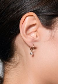 Złota kolczyk w kształcie obręczy z zawieszką w formie kropli, z przezroczystym kamieniem szlachetnym, o gładkim wykończeniu i lekkiej konstrukcji, noszony na płatku ucha.