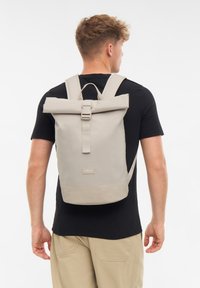 Man with short hair wearing black t-shirt and beige pants, carrying a beige roll-top backpack with buckle on his back.