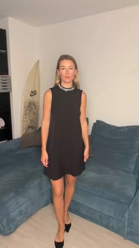 Fekete ujjatlan ruha ékszerrel díszített nyakkivágással, laza szabással és oldalsó sliccel, fekete magassarkú cipővel, egy texturált kék kanapén.