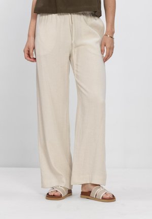 Vrouw draagt losse beige broek en beige sandalen met bruine zolen, staande tegen een effen lichte achtergrond.