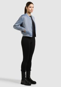 Femme debout portant une veste bomber en satin bleu clair, un haut noir, des leggings noirs et des bottes noires mi-mollet sur un fond uni.
